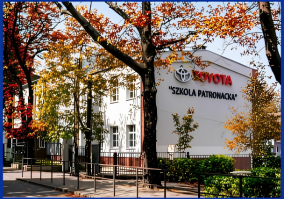 Biały budynek Zespołu Szkół nr 1 w Rudzie Śląskiej otoczony drzewami z jesiennymi, żółto-czerwonymi liśćmi. Na bocznej elewacji szkoły widoczne jest duże, czerwone logo oraz napis „TOYOTA SZKOŁA PATRONACKA”. Przed budynkiem znajduje się metalowe ogrodzenie oraz chodnik
